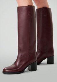 Bottes en cuir bordeaux jusqu'au genou avec une finition lisse, un talon carré et une couture décorative le long des coutures latérales.