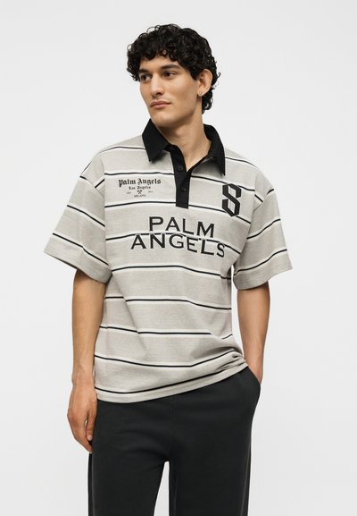 Νέος με σγουρά μαλλιά φοράει γκρι ριγέ πόλο μπλούζα με μαύρο γιακά και το κείμενο «Palm Angels», συνδυασμένη με μαύρο παντελόνι, στέκεται μπροστά σε λευκό φόντο.