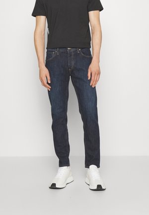 Jeans de mezclilla azul oscuro con cinco bolsillos, botón de metal y corte estándar, combinados con una camiseta negra de manga corta y zapatillas blancas.