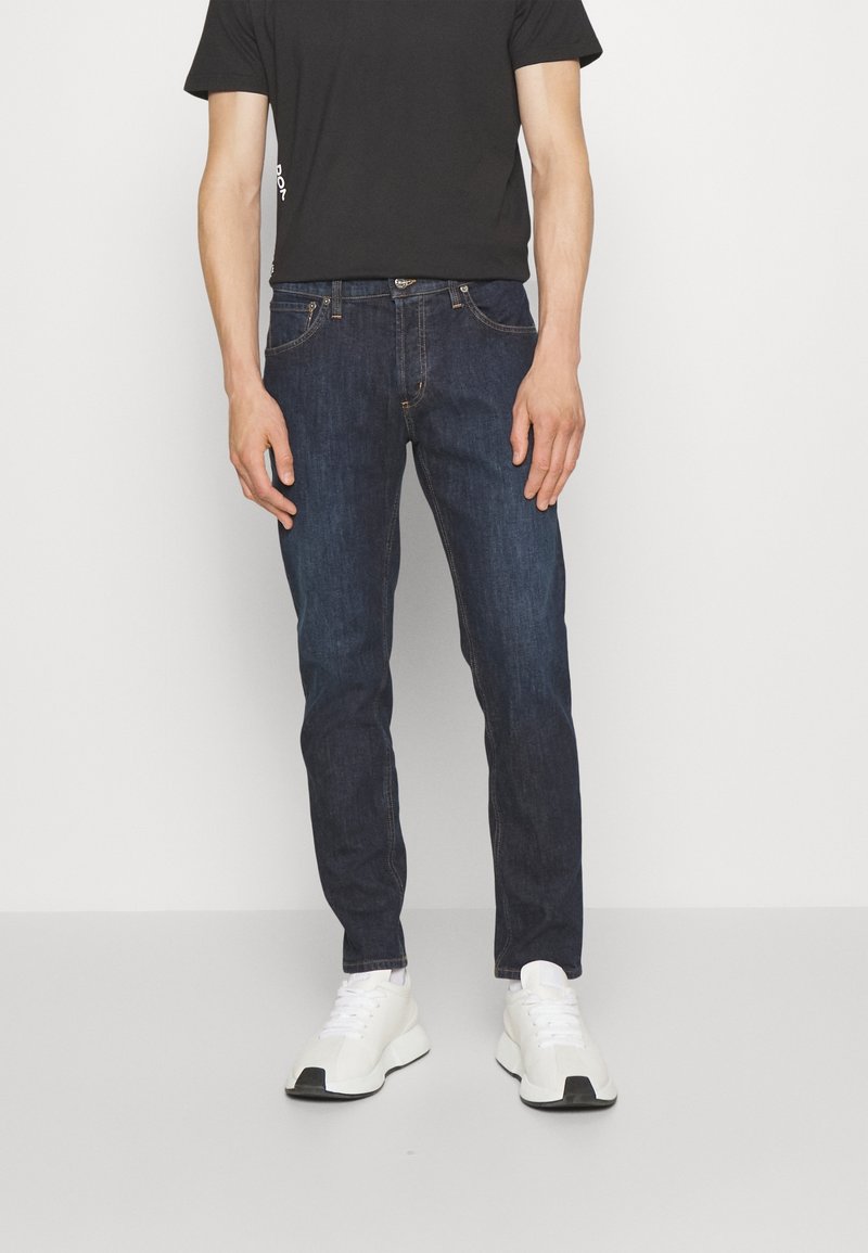 Granatowe dżinsy z pięcioma kieszeniami, metalowym guzikiem i standardowym krojem, noszone z czarną koszulą z krótkim rękawem i białymi trampkami.