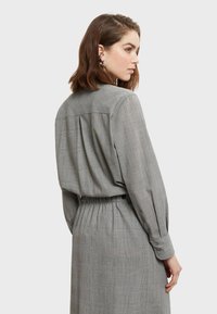 Siva karičasta haljina s dugim rukavima, dugmadi na manžetama i suženim strukom. Ima nabor na leđima za dodatni volumen i teksturu.