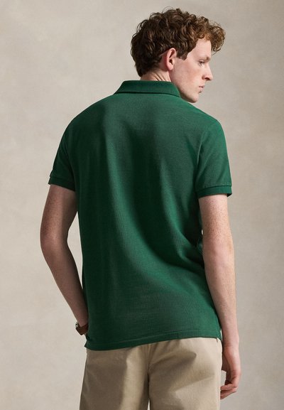 Νεαρός άνδρας με σγουρά κόκκινα μαλλιά, φορώντας σκούρο πράσινο πόλο με κοντά μανίκια και μπεζ σορτς, στραμμένος πλευρικά σε απλό φόντο.