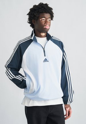 Hellblaues und marineblaues, zum Zuziehen geeignetes Trainingsjacke mit drei weißen Streifen an den Ärmeln, Adidas-Logo auf der Brust und gerippten Bündchen. Weiches, leichtes Material.