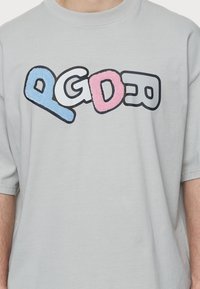 Camiseta de manga corta en gris claro con letras texturizadas y coloridas "PGD" en azul, rosa y negro. Diseño sencillo y de corte relajado.