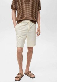 Lichtbeige shorts met een elastische tailleband en trekkoord. Draag een bruin gestreept gebreid T-shirt met korte mouwen en bruin instap-sandalen.