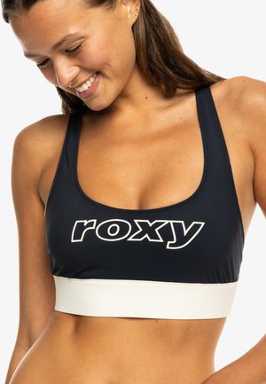 Lachende vrouw die naar beneden kijkt in een zwarte sportbeha met het witte "roxy" logo en een brede witte band eronder.