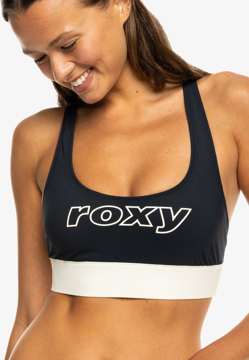 Usmievajúca sa žena pozerajúca dolu, oblečená v čiernom športovom podprsenke s bielym logom "roxy" a širokým bielym pásom pod ním.