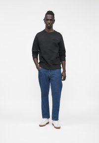 Černá mikina s texturovaným vzorem, v kombinaci se světle modrými džínami a bílými teniskami s hnědými podrážkami, stojící na jednobarevném pozadí.