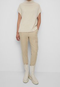 Pull en maille beige avec col haut et ourlet en côtes, associé à un pantalon cargo beige avec poches latérales et poignets. Chaussures : bottes beiges.
