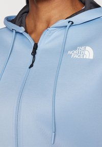 Světle modrá mikina na zip s šedým vnitřkem, má černý zip, šňůrky a bílý logo na levé části hrudi.