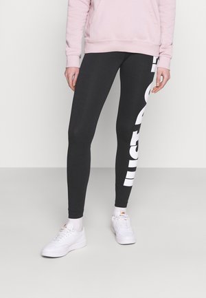 Leggings noirs avec une coupe ajustée, arborant un grand logo blanc sur le côté gauche ; associés à un sweat-shirt rose clair et à des chaussures blanches.