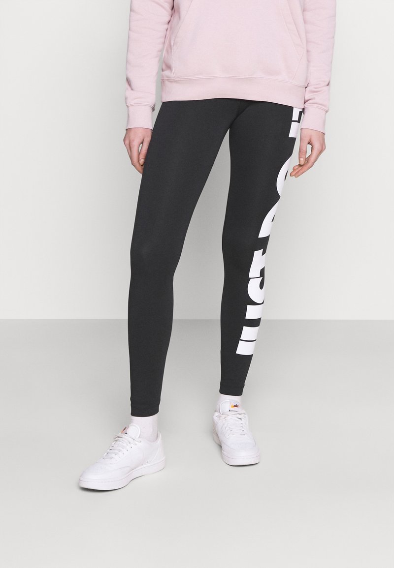 Svarta leggings med en figurnära design, prydda med en stor vit logotyp på vänster sida; tillsammans med en ljusrosa sweatshirt och vita skor.