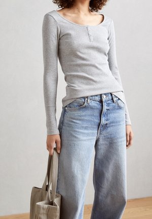 Personne portant un haut gris clair à manches longues et un jean bleu ample, tenant un tote bag beige, debout en intérieur contre un mur uni.