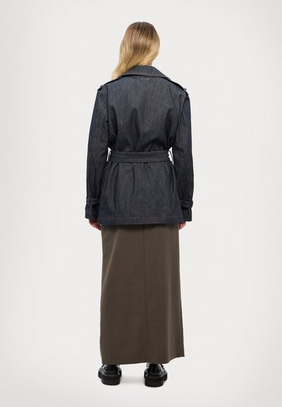 Veste en denim foncé avec une taille ceinturée, un col classique et des manches longues, associée à une longue jupe olive et des chaussures à plateforme noires.