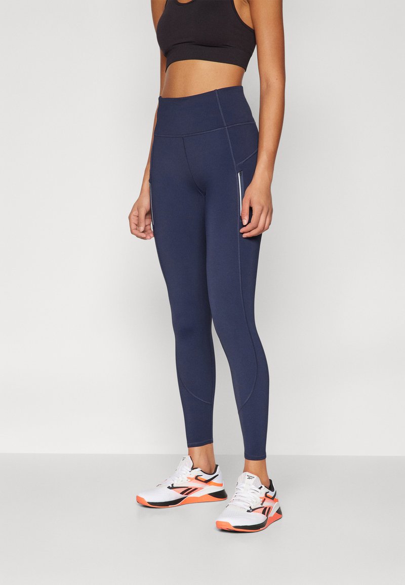 Marineblå høyt liv leggings med tonede sømmer og sidelommer med glidelås, sammen med hvite og oransje treningssko. Glatt, elastisk stoff.