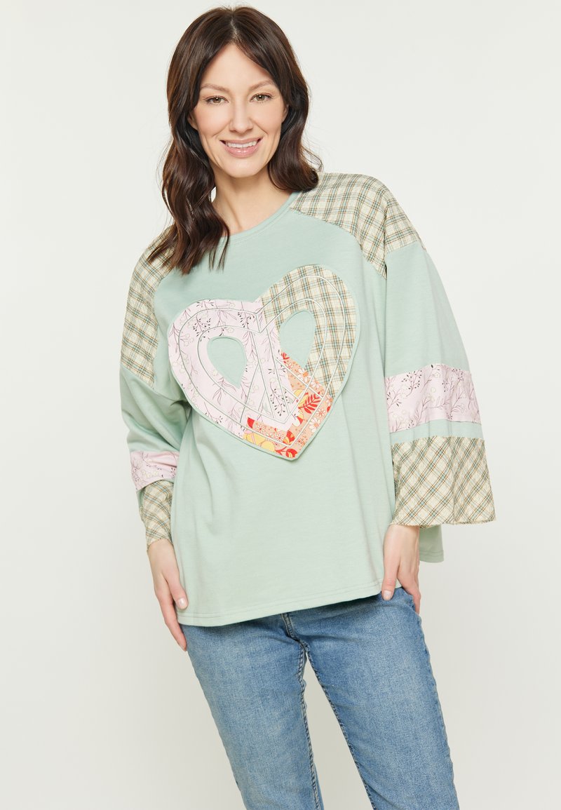 Világoszöld, nagyméretű pulóver, nagy szív alakú folttal, virágos és kockás mintával az ujjain és díszítő részleteken.