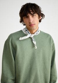 Green sweatshirt with a crew neck, soft texture, and long sleeves. Patterned white scarf tied at the neck adds contrast.