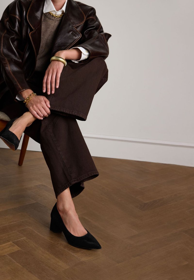 Pantaloni larghi marroni abbinati a una giacca di pelle color cioccolato. Décolleté in suede neri con tacco a blocco e dettagli in oro ai polsi.