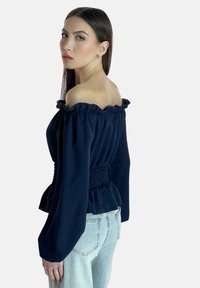 Marineblaues Off-Shoulder-Oberteil mit gerafftem Ausschnitt, gesmokter Taille und langen Ärmeln, kombiniert mit hellblauen Jeans. Glatter Stoff und tailliertes Design.