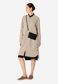 Beżowy płaszcz z mieszanki wełny z zamkiem z przodu, bocznymi guzikami i asymetrycznym dołem. W połączeniu z czarną torbą na ramię oraz brązowymi mokasynami.