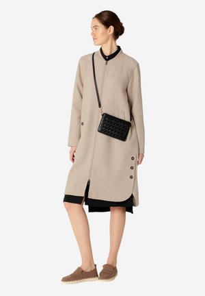 Cappotto beige in misto lana con zip frontale, bottoni laterali e orlo asimmetrico. Abbinato a una borsa a tracolla nera e mocassini marroni.