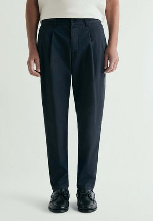 Hombre con pantalones negros a medida con pliegues y zapatos de cuero negros con cordones, de pie frente a un fondo claro y liso.