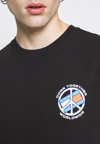 Juodas medvilnės marškinėliai su apvalia iškirpte, puošti spalvingu apskrito taikos laikrodis grafiku ir tekstu: "ATEIKITE KARTU VISAME PASAULYJE."