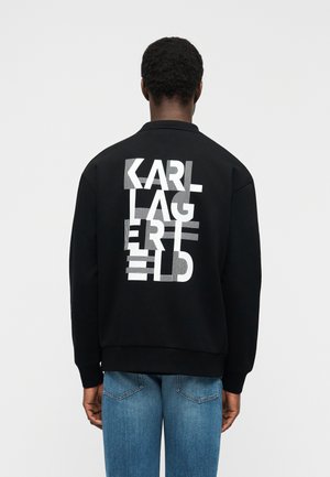 Persona che indossa una felpa nera con il testo bianco distorto "KARL LAGERFELD" sul retro, abbinata a jeans blu, di spalle.