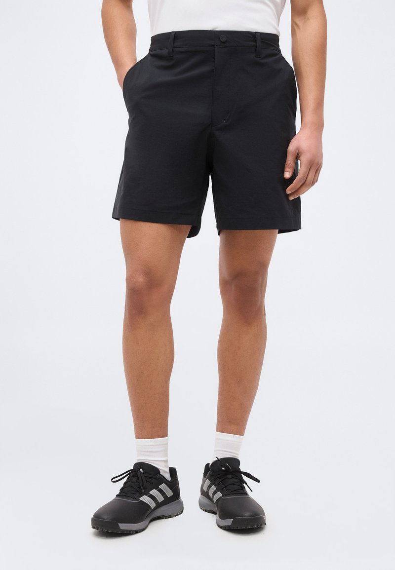 Άνδρας που φοράει μαύρο σορτς, λευκές κάλτσες και μαύρα αθλητικά παπούτσια με λευκές ρίγες, στέκεται με τα χέρια κοντά στις τσέπες.