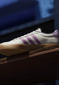 Zapatillas blancas y de un púrpura claro con tres franjas paralelas, parte superior de cuero texturizado, puntera redonda y suela de goma con un patrón.