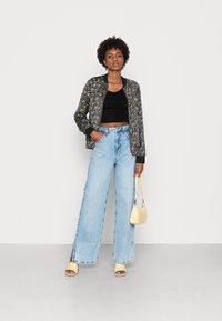 Jaqueta bomber preta com flores sobre um top crop preto, combinada com jeans de perna larga azul claro e sandálias de salto amarelas, segurando uma pequena bolsa bege.