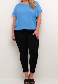 Femme portant un haut bleu ample, un pantalon noir ajusté et des chaussures à talons noires ouvertes à l'avant, se tenant devant un fond blanc uni.