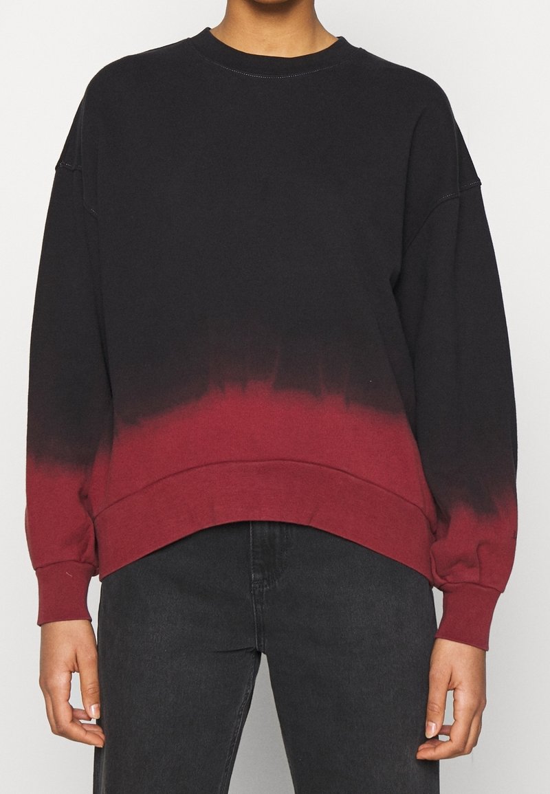 Sweat-shirt à manches longues dégradé noir et rouge avec col rond, porté avec un jean foncé, visible du milieu de la poitrine jusqu'aux hanches.