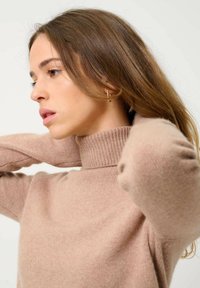 Beige Rollkragenpullover mit geripptem Kragen, weichem Material und langen Ärmeln. Das Model zeigt eine entspannte Haltung und gestylte Haare.
