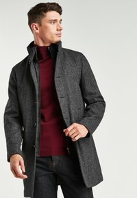 Grau strukturierter Mantel mit Fischgrätmuster, Druckknopfverschluss und hohem Kragen über einem roten Rollkragenpullover, kombiniert mit dunklen Jeans.