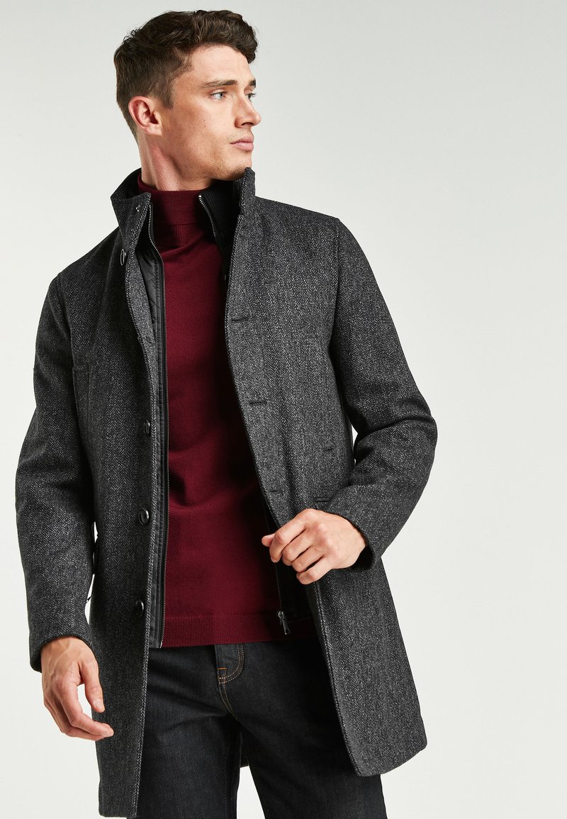 Grau strukturierter Mantel mit Fischgrätmuster, Druckknopfverschluss und hohem Kragen über einem roten Rollkragenpullover, kombiniert mit dunklen Jeans.