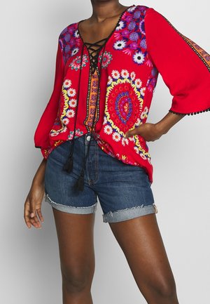 Femme portant un chemisier rouge à motif floral avec un laçage noir à l'avant et des manches trois-quarts, associé à un short en jean retroussé.