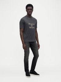 Млад мъж, облечен с сива тениска Emporio Armani, черни дънки и черни маратонки с бели подметки на едноцветен светъл фон.