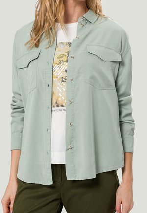 Frau trägt ein salbeigrünes, geknöpftes Hemd über einem weißen T-Shirt mit Blumenmuster und dunkelgrüne Hose, Arme entspannt seitlich am Körper.