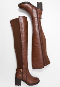 Botas marrones por encima de la rodilla con acabado en cuero liso, laterales de tela elástica, tacón bloque y cremallera lateral. Detalle de hebilla en el tobillo.