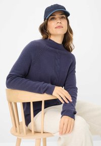 Navyblauw gebreid sweater met hoge nek en kabelpatroon, gedragen met crèmekleurige loungebroek, zittend op een houten stoel.