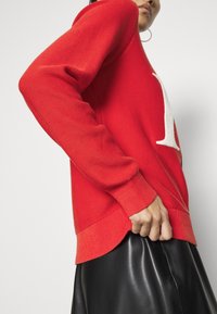 Femme portant un pull rouge en maille avec un détail de lettre blanche et une jupe en cuir noir, posant contre un fond uni clair.