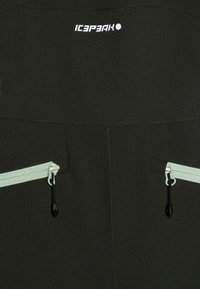 Mørk grønn softshell-jakke med frontlogo, to glidelåslommer med mintgrønne glidelåser, og en glatt tekstur.