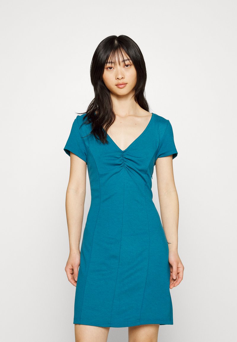 Vestido justo em azul-petróleo em tecido suave, com decote em V, mangas curtas e detalhes franzidos na área do peito. Apresenta uma saia levemente evasé.