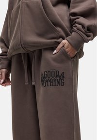 Bruine hoodie en joggingsbroek van zacht materiaal. De joggingsbroek heeft een zwart grafisch logo op de dij, met zichtbare trekkoorden en zakken.