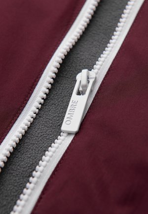 Cremallera blanca con la marca "OMBRE" parcialmente abierta en una chaqueta granate, revelando un forro de vellón gris oscuro debajo.