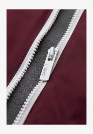 Weißer Reißverschluss mit "OMBRE"-Logo, teilweise geöffnet an einer bordeauxroten Jacke, die ein dunkelgraues Fleece-Futter darunter zeigt.