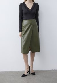Top nero a maniche lunghe con scollatura in pizzo, abbinato a una gonna avvolgente in ecopelle verde. L'insieme è completato da scarpe nere con tacco alto.