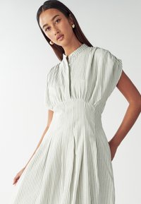 Svetle zelené šaty s bílými pruhy, krátkými nabíranými rukávy, zapínáním na knoflík a skládáním sukně, která elegantně vlaje.