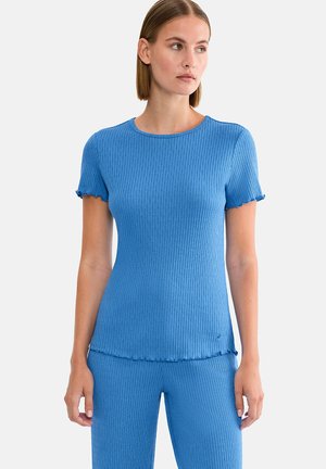 Femme portant un haut bleu côtelé à manches courtes ajusté et un pantalon assorti, debout avec les bras détendus, regardant sur le côté sur un fond blanc.